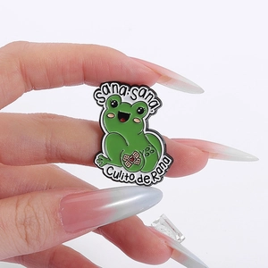 Detail of cute green frog enamel pin with Sana Sana text held by hand on white background