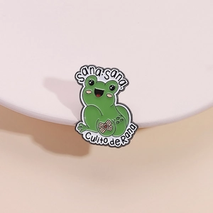 Cartoon green frog enamel pin with Sana Sana Culito de Rana text on a display stand