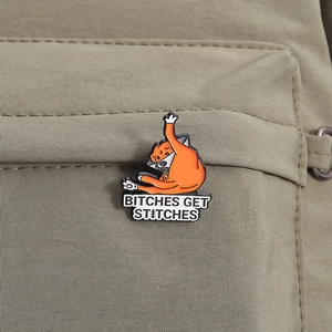 Close-up of orange cat enamel pin with Bitches Get Stitches text on olive green fabric