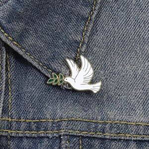 Detail view of a white dove enamel pin with a green olive branch on denim fabric