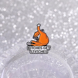 Funny orange cat enamel pin with Bitches Get Stitches quote on a glass rim
