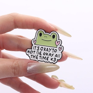 Nahaufnahme einer grünen Frosch-Emaille-Anstecknadel mit Text, in der Hand gehalten