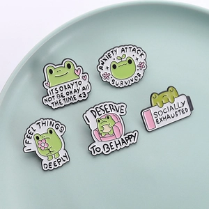 Set aus fünf niedlichen grünen Emaille-Frosch-Pins mit Sprüchen auf einem Keramikteller
