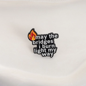 Enamel pin with orange flame and quote may the bridges i burn light my way on cream base
