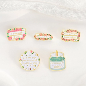 Five motivational enamel pins with floral designs and golden borders on white fabric