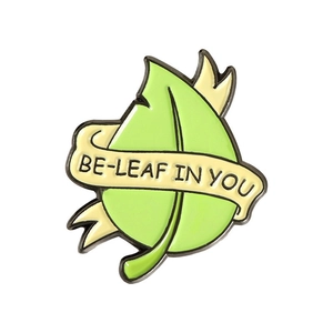 Close-up of leaf enamel pin with BE-LEAF IN YOU quote and yellow ribbon detail