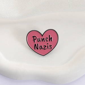Macro view of heart-shaped pink enamel pin with black Punch Nazis text on white stand