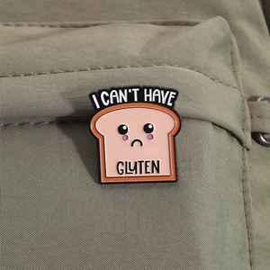 Gros plan d'un pin's en émail toast rose avec 'I CAN'T HAVE GLUTEN' sur un sac vert