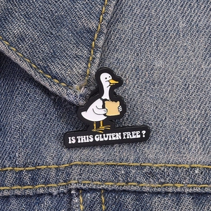 Cartoon white duck holding bread enamel pin with Is This Gluten Free slogan on denim