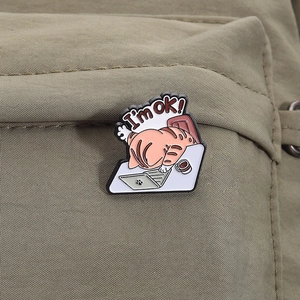 Detailed close-up of a funny 'I'm ok!' cat enamel pin on a dark green canvas bag