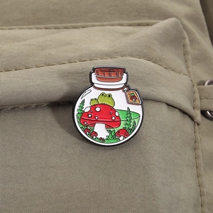 Close-up of frog and mushroom terrarium enamel pin on green canvas backpack strap