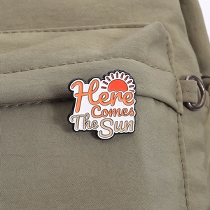 Detail of a 'Here Comes The Sun' sun enamel pin attached to a green nylon fabric bag