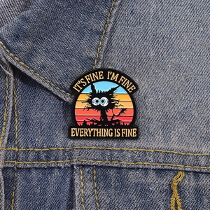 Rainbow cat enamel pin with It's Fine meme text on dark blue denim jacket fabric
