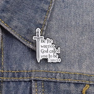 Close-up of sword enamel pin with 1 John 5:4 quote on blue denim jacket collar