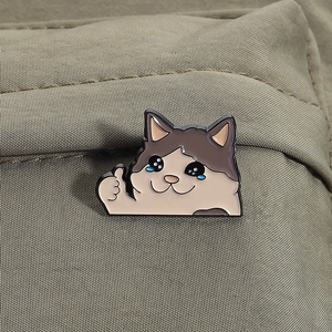 Gros plan du pin's émaillé chat qui pleure pouce levé sur tissu vert olive