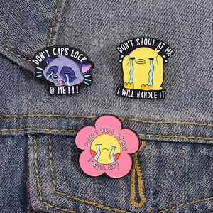 Three cute crying animal enamel pins on denim, including a duck, raccoon, and flower