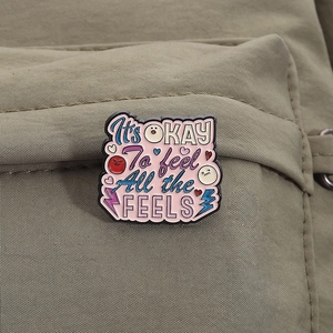 Nahaufnahme der rosa 'It's OK' Emaille-Anstecknadel mit Herz-Symbol auf grünem Segeltuch