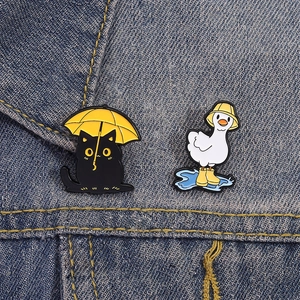 Cute enamel pins featuring a black cat with an umbrella and a duck on blue denim fabric