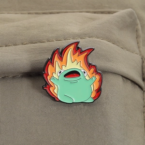 Detailaufnahme einer grünen Frosch-Emaille-Nadel mit bunten Flammen auf schwarzer Tasche