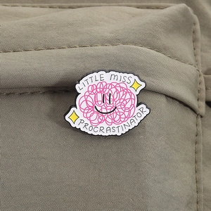Close-up of pink yarn ball smile brooch with Little Miss Procrastinator text on green bag