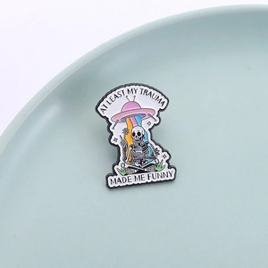 Skeleton and UFO enamel pin featuring At Least My Trauma Made Me Funny text on ceramic