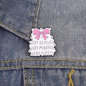 Cute white enamel pin with pink bow and 'Very Demure' quote on denim jacket