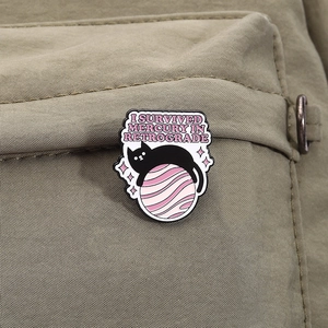 Detail of I Survived Mercury Retrograde enamel pin with a black cat on a pink planet