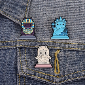Three creative cat enamel pins featuring world landmarks on blue denim fabric
