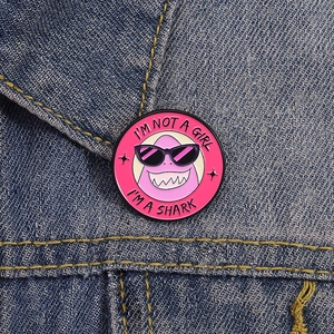 Pink round enamel shark pin with I'm Not a Girl text on blue denim jacket