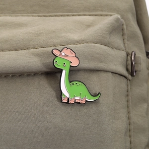 Close-up of a green dinosaur enamel pin with pink cowboy hat on olive green canvas bag