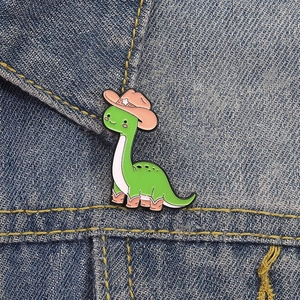 Creative cartoon green dinosaur enamel pin wearing a pink cowboy hat on denim background