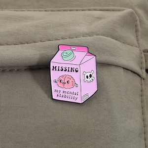 Detail der rosa Emaille-Brosche in Milchkarton-Form mit humorvollem Text auf Stoffoberfläche