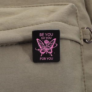 Close-up of black enamel butterfly brooch with Be You pink text on green fabric