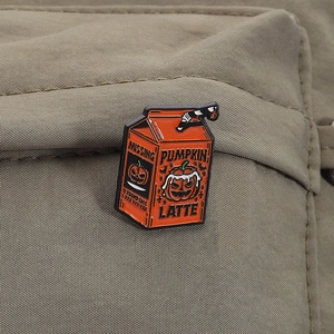Detail of pumpkin latte milk box enamel pin pinned on green canvas fabric background