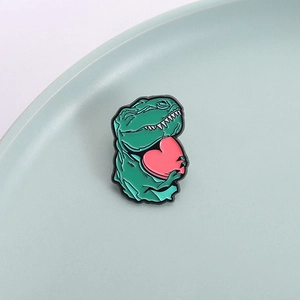 Cute green dinosaur enamel pin holding a pink heart on a light ceramic tray