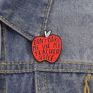 Close-up of a teacher-themed red apple enamel pin on a blue denim jacket collar