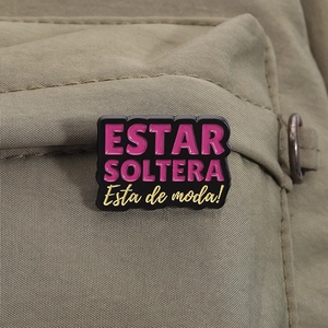 Detail of Estar Soltera enamel pin with pink and yellow text on a fabric bag