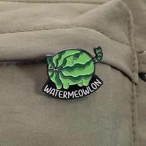 Detailed close-up of a watermelon cat enamel pin on green fabric texture