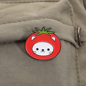Detail of a cute cartoon white cat enamel pin with a red tomato hat on green fabric