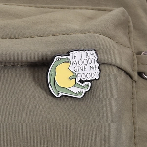Funny green frog enamel pin with 'If I am moody give me foody' text on olive backpack