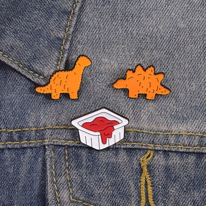 Cartoon dinosaur chicken nugget and tomato sauce enamel pins set on denim fabric