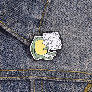 Funny green frog enamel pin with 'If I am moody give me foody' text on denim background