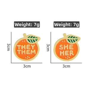 Détail des épinglettes en émail orange avec lettrage des pronoms They/Them et She/Her