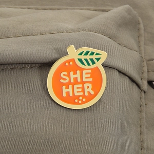 Détail d'un pin's en émail en forme d'orange avec pronom SHE HER sur une veste en tissu