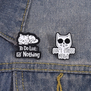 Détail de pin's chat en émail sur denim, montrant le texte 'To Do List: Nothing' en gros plan