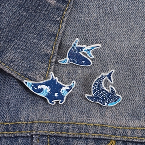 Close-up of three ocean-themed enamel pins featuring shark and ray on denim fabric