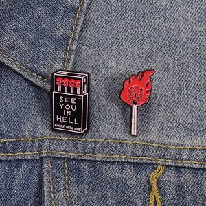 Macro detail of gothic See You In Hell matchbox and skull flame enamel pins on denim fabric