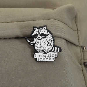 Detail showing cartoon raccoon enamel pin with i require snacks text on a green canvas bag