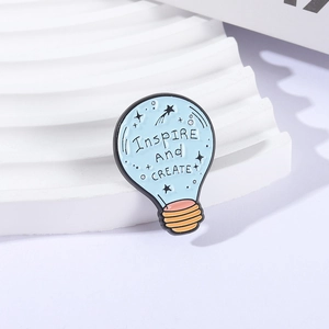 Light blue lightbulb enamel pin with Inspire And CREATE slogan on a white pedestal