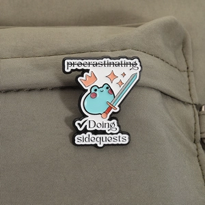 Enamel pin featuring a blue frog with a sword on a khaki backpack with a sidequest slogan